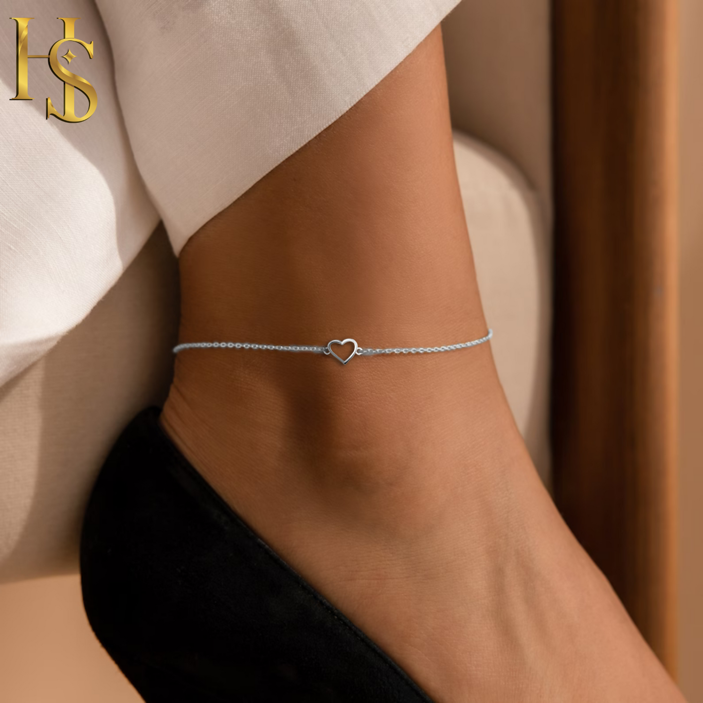 A delicate sterling silver anklet with a central open heart charm, worn on the ankle above a black shoe.