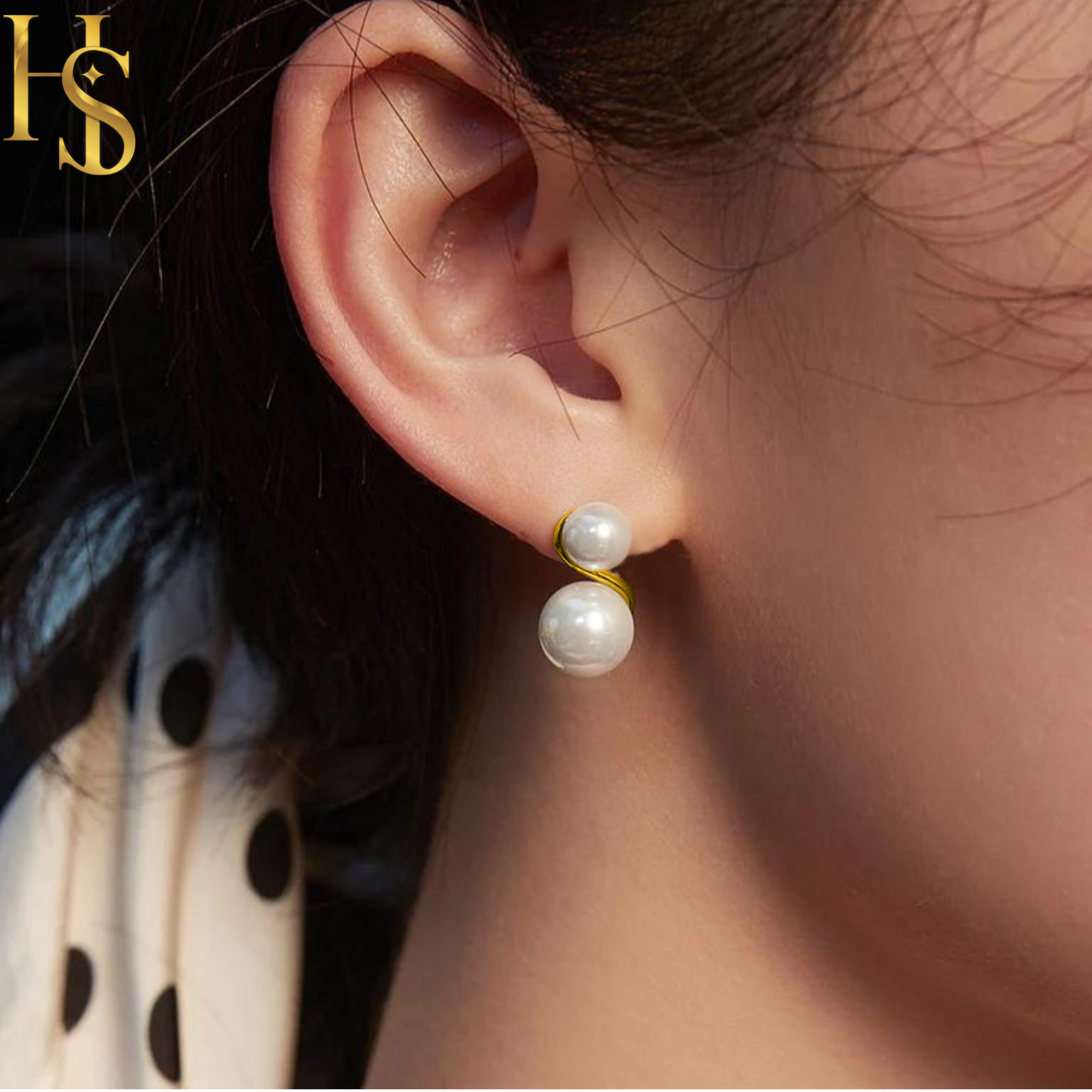 Gold-plated stud earrings with two white pearls in a spiral setting, worn on the ear with a polka dot scarf in the background.
