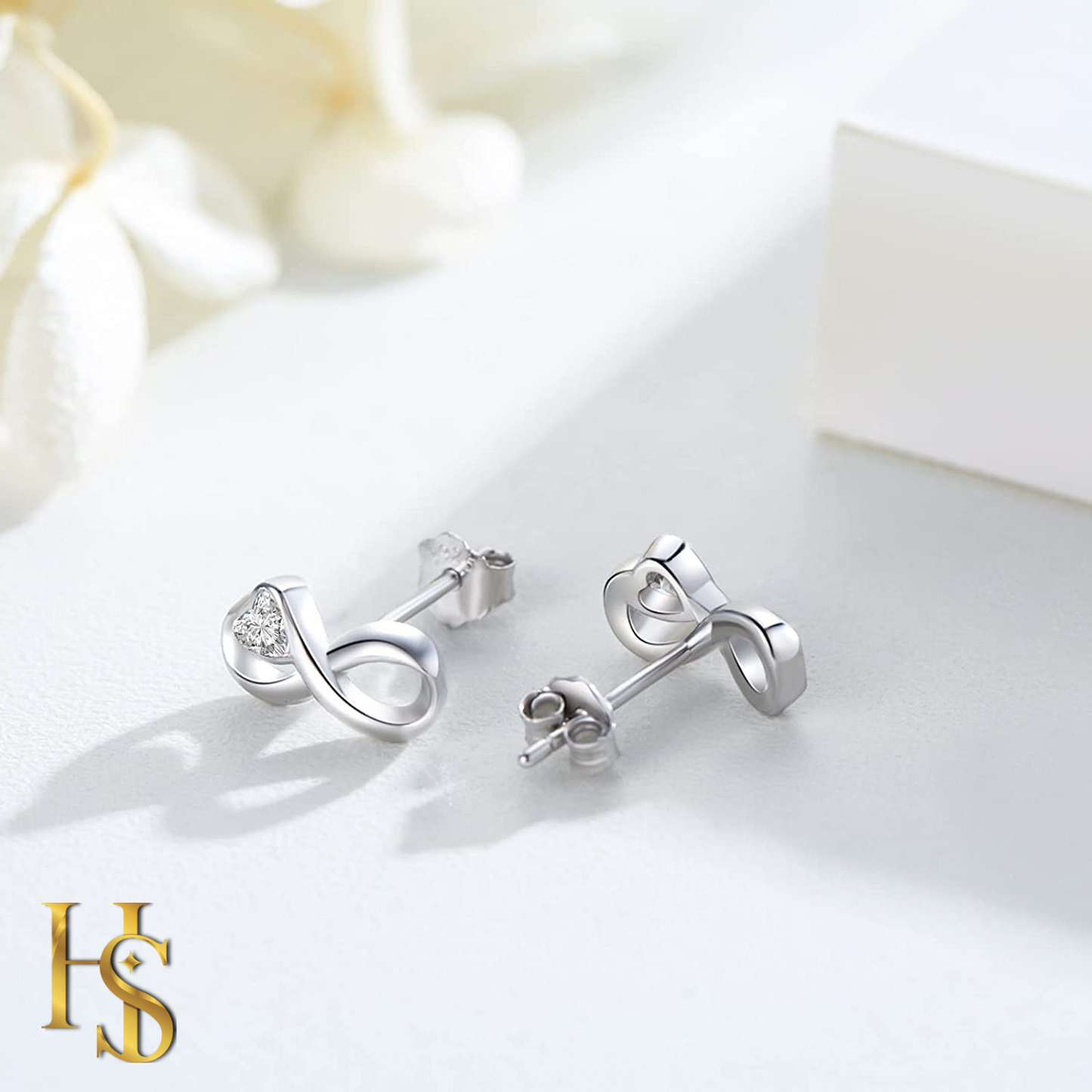 A pair of infinity-shaped stud earrings in sterling silver with a single American diamond, styled on a white surface with soft flowers.