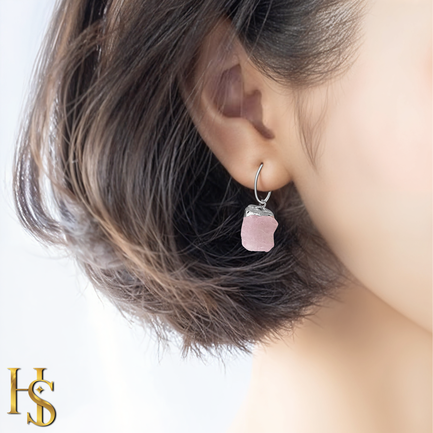 A sterling silver hoop earring with a dangling raw, rough-cut rose quartz gemstone is worn on the ear.