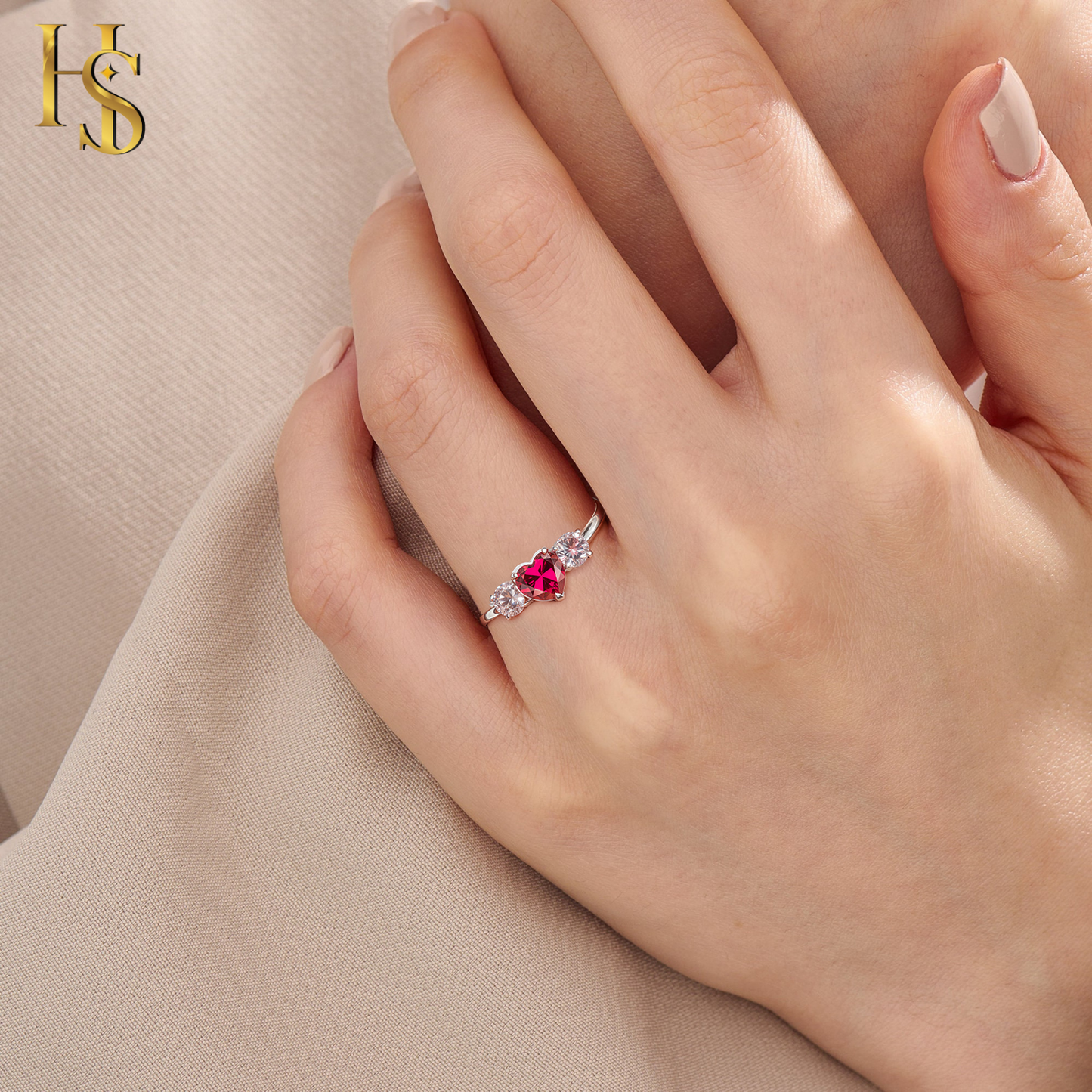 A sterling silver adjustable ring with a central heart-shaped red stone and two side American diamonds worn on a hand against beige fabric.