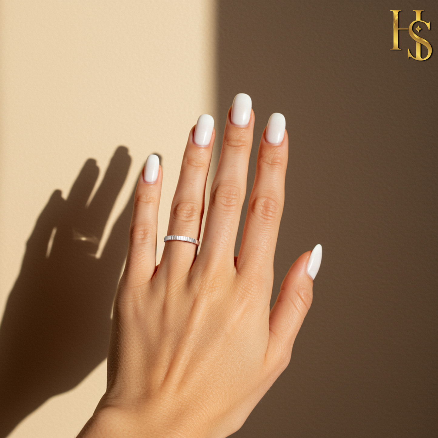 A simple textured sterling silver band ring worn on a hand with white nails, set against a beige background with a strong shadow.