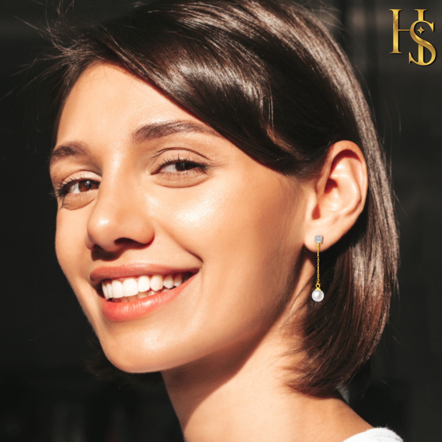 A smiling woman wears gold-plated drop earrings with a pave American diamond stud and a dangling pearl, seen in warm sunlight.