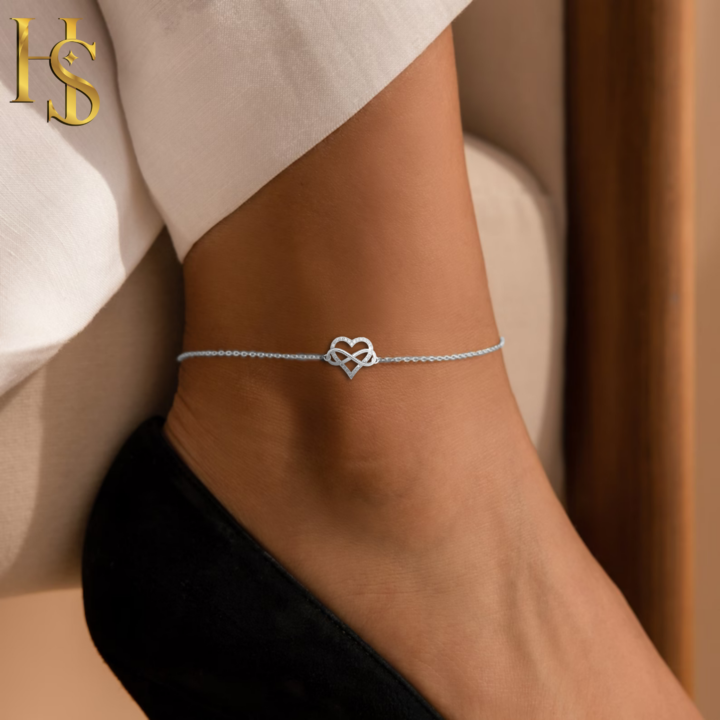 A sterling silver anklet with a heart and infinity motif, studded with tiny American diamonds, worn on the ankle with a black shoe.