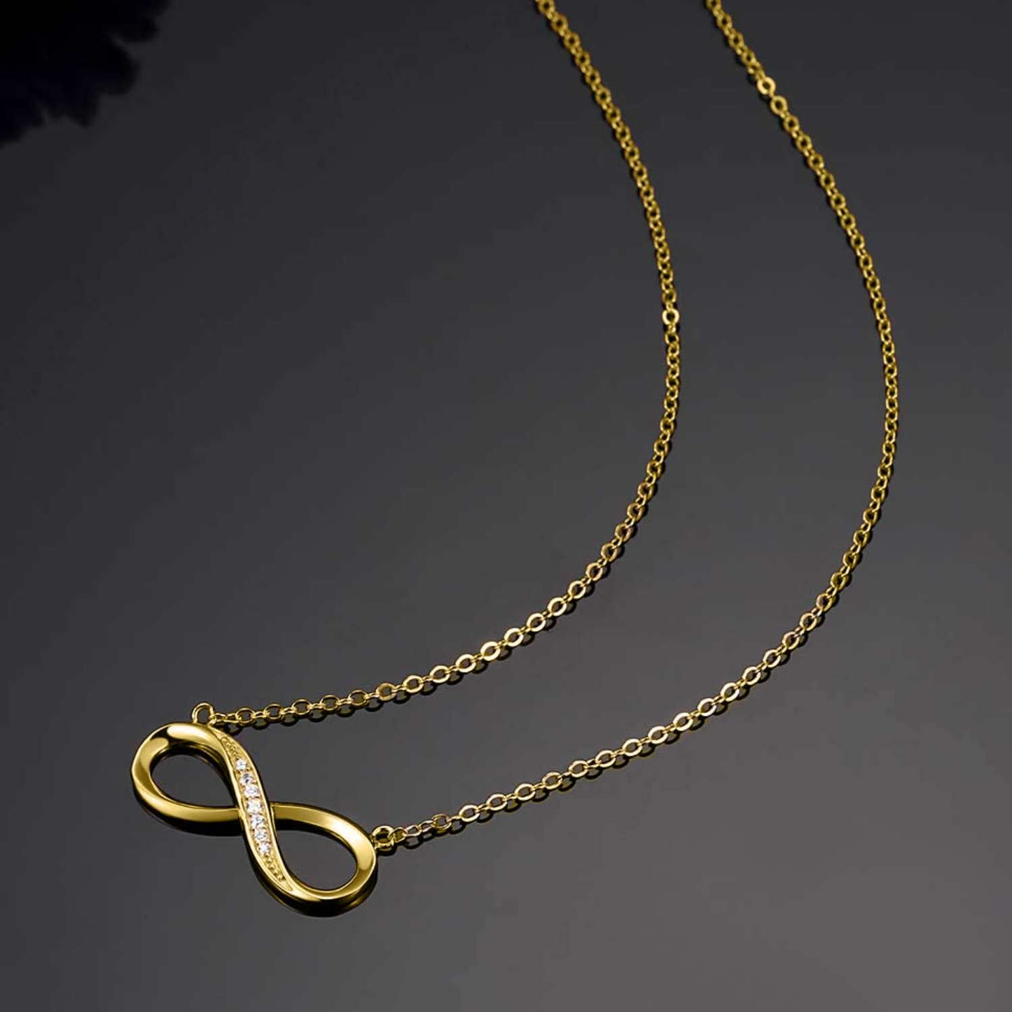 A gold-plated infinity pendant necklace with pavé-set American diamonds on one curve, styled on a dark reflective surface.