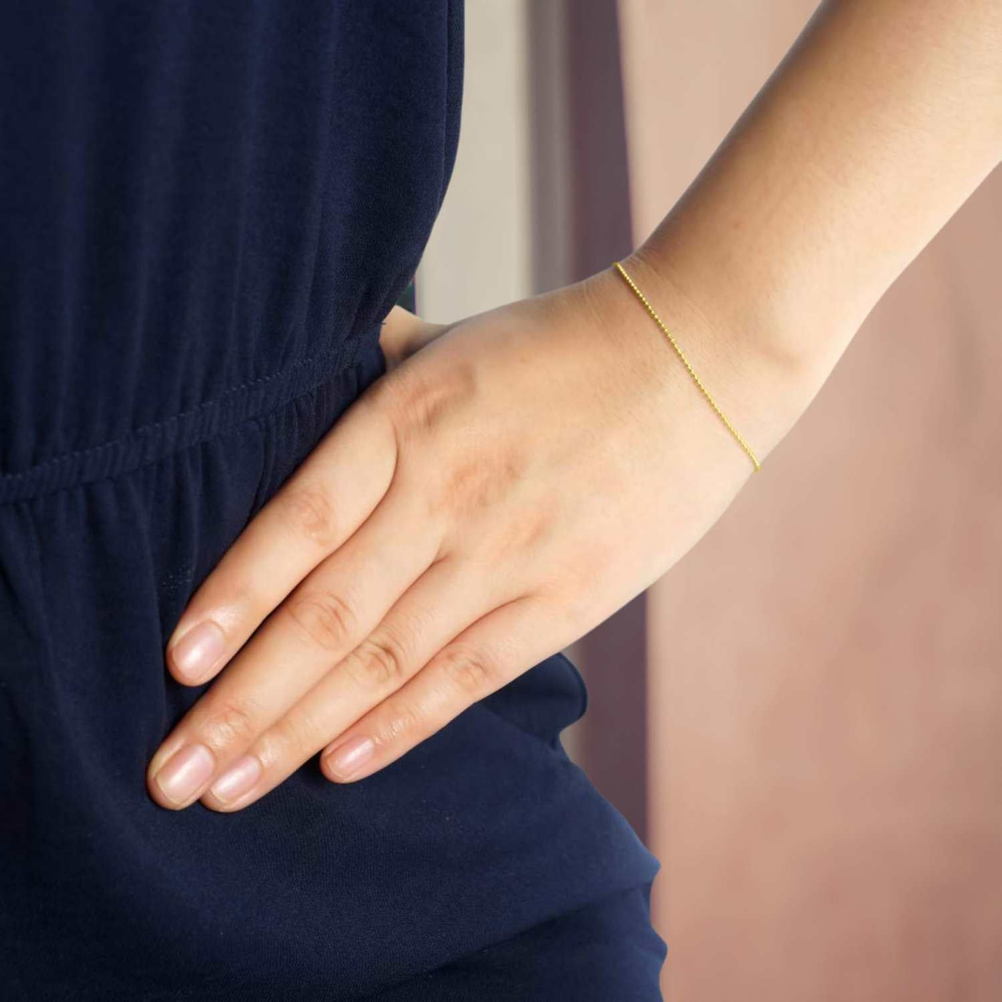 A delicate gold-plated ball chain bracelet worn on the wrist of a model with her hand on her hip.
