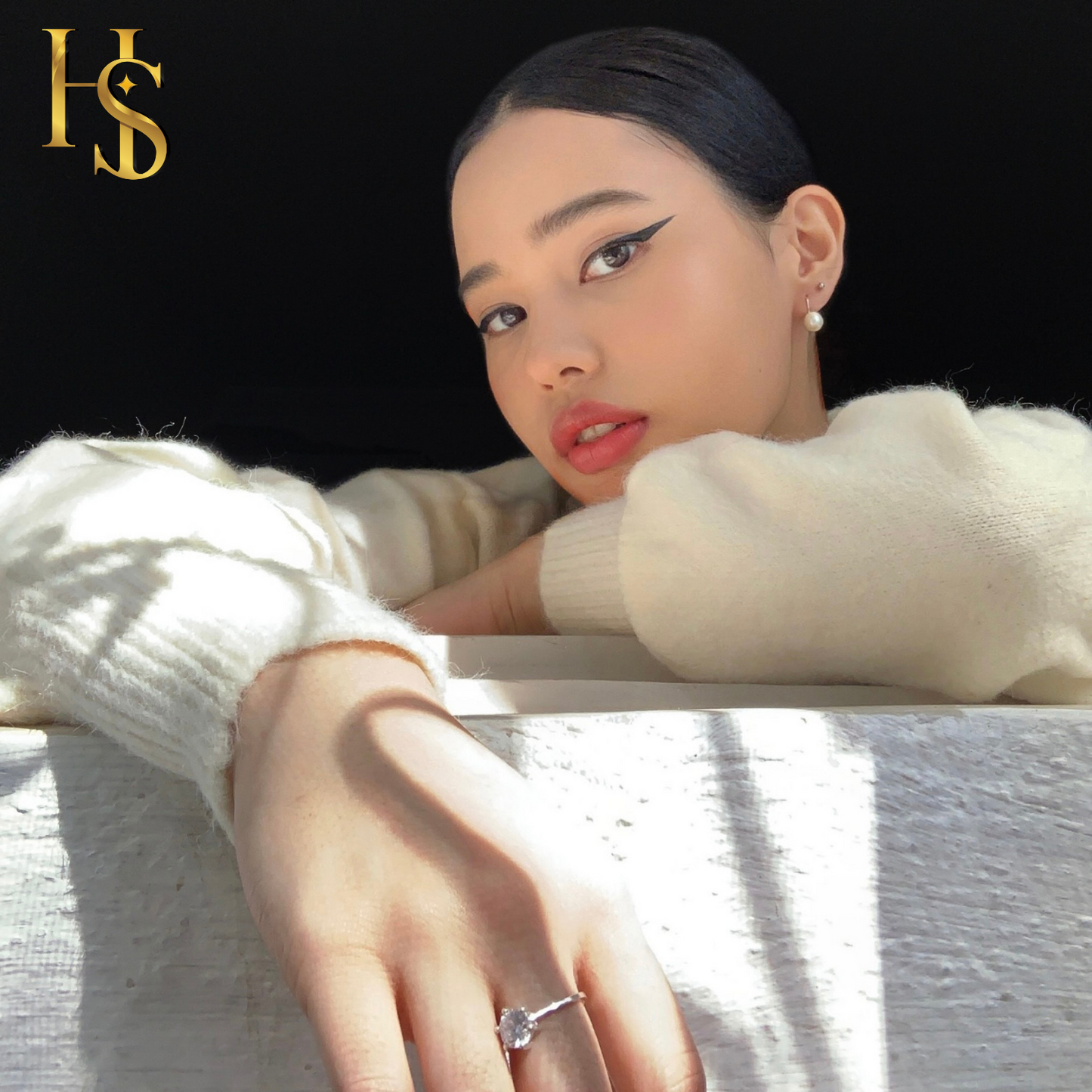 A woman wears a solitaire ring with a round American diamond in a four-prong setting, resting her hand on a white ledge in the sun.