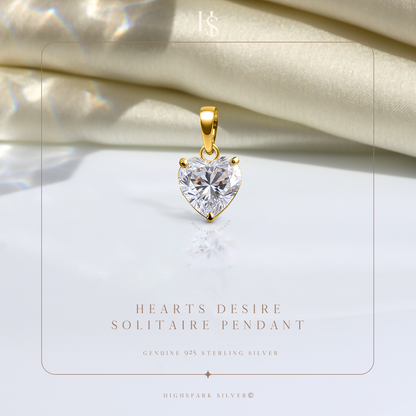 A heart-shaped solitaire American diamond pendant with a three-prong setting and gold-plated bail, styled on a reflective white surface.
