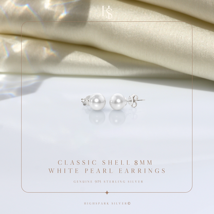 A pair of classic pearl stud earrings in sterling silver with push-back closures, styled on a reflective surface with draped cream satin.