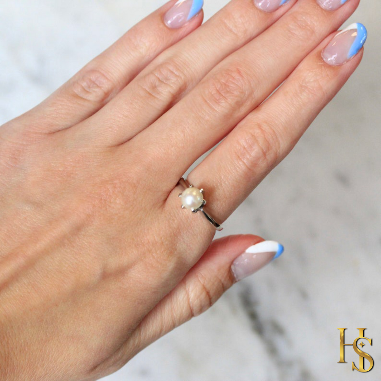 A solitaire pearl ring with a six-prong setting and a sterling silver band, worn on a hand with a blue and white manicure.