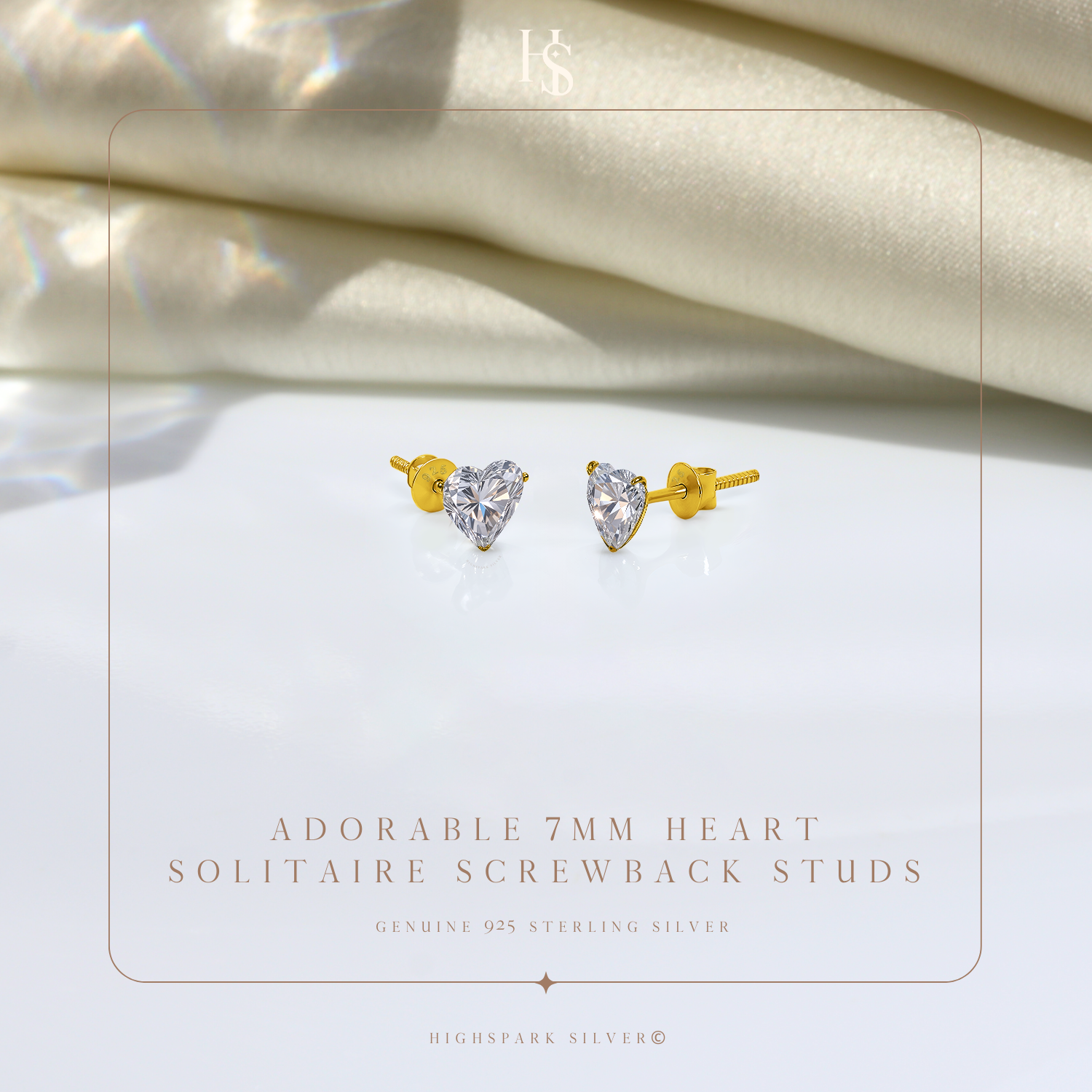 A pair of gold-plated stud earrings with heart-shaped American diamonds and screw backs, styled on a white surface with draped satin.