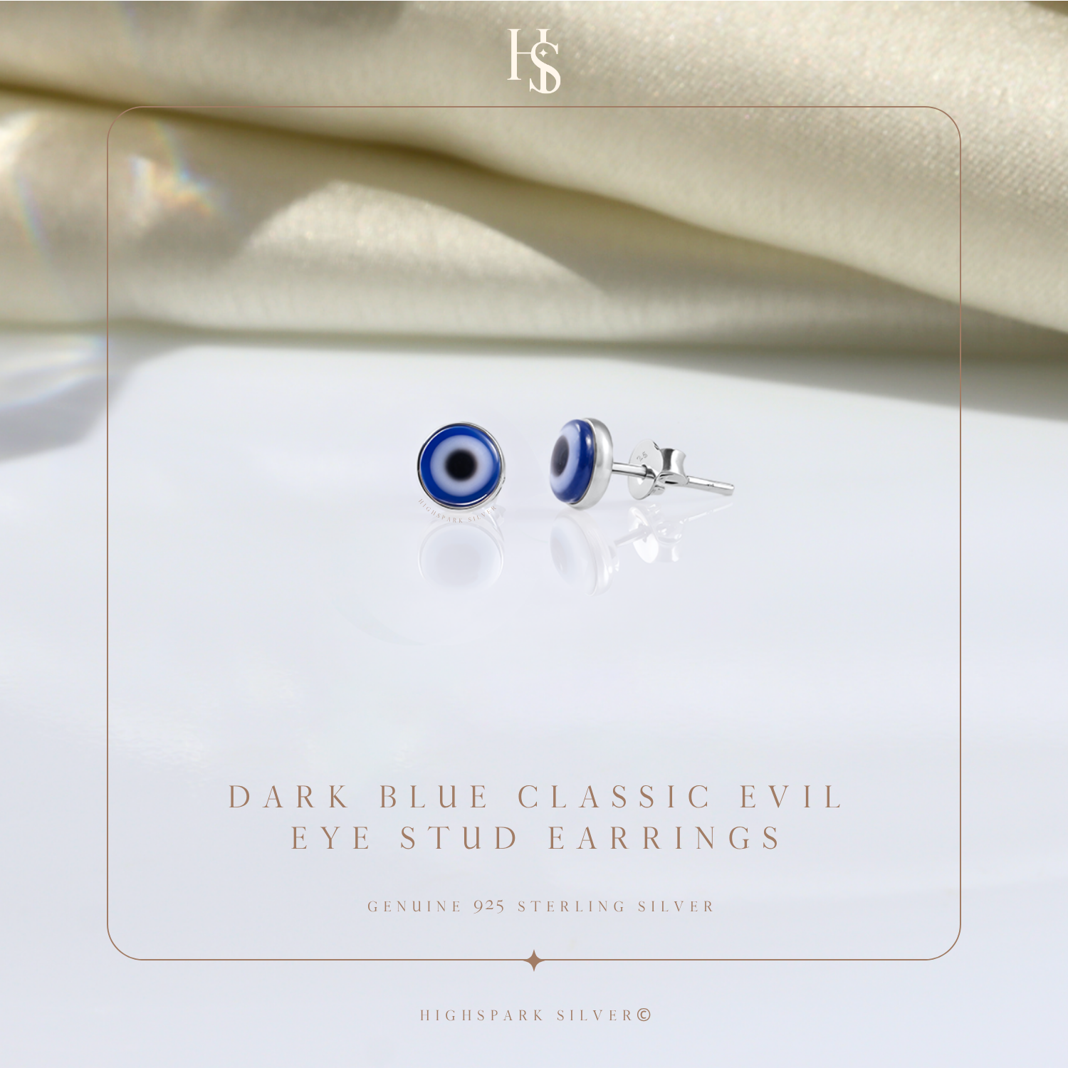 A pair of round evil eye stud earrings in sterling silver, styled on a reflective surface with draped cream fabric in the background.