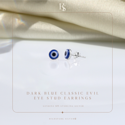 A pair of round evil eye stud earrings in sterling silver, styled on a reflective surface with draped cream fabric in the background.
