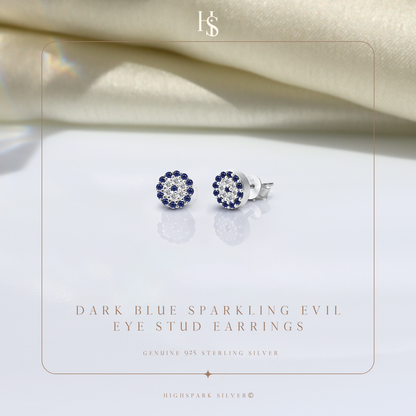 A pair of evil eye stud earrings with blue and clear American diamonds in sterling silver, styled on a reflective surface with satin fabric.