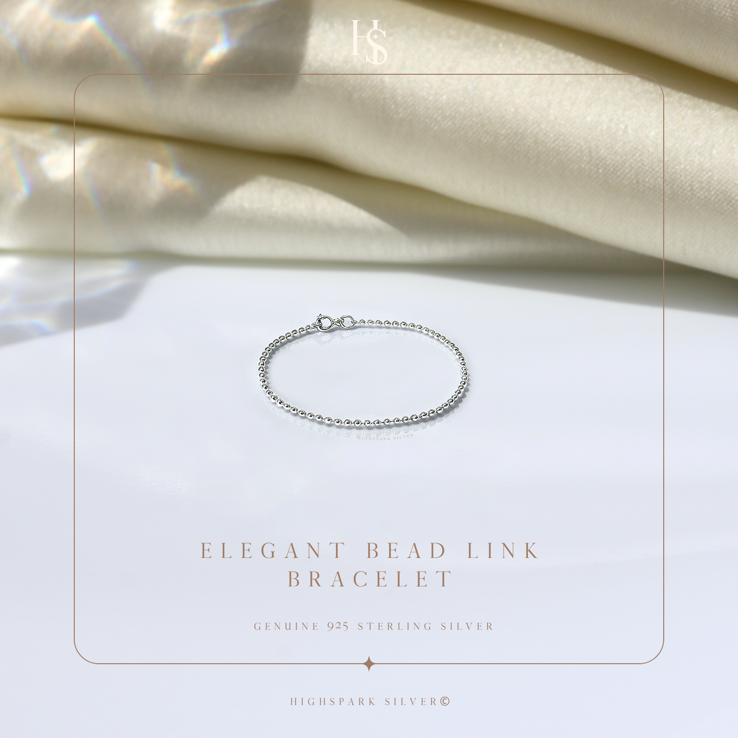 A delicate sterling silver bead link bracelet with a spring ring clasp, styled on a white surface with draped cream satin fabric.