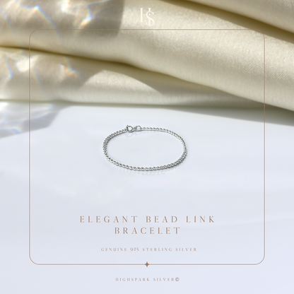 A delicate sterling silver bead link bracelet with a spring ring clasp, styled on a white surface with draped cream satin fabric.