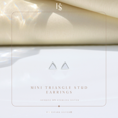 A pair of mini triangle stud earrings in sterling silver, styled on a white surface with a soft beige fabric background.