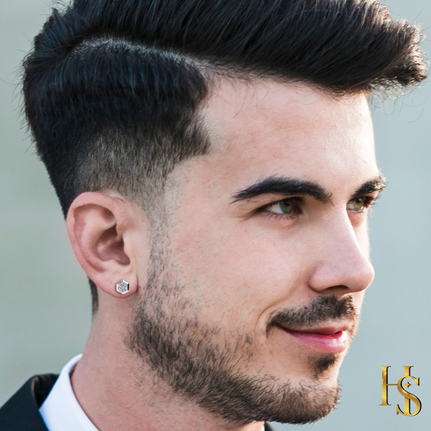 A hexagonal solitaire stud earring with a central American diamond in a sterling silver bezel setting, worn on a man's ear.