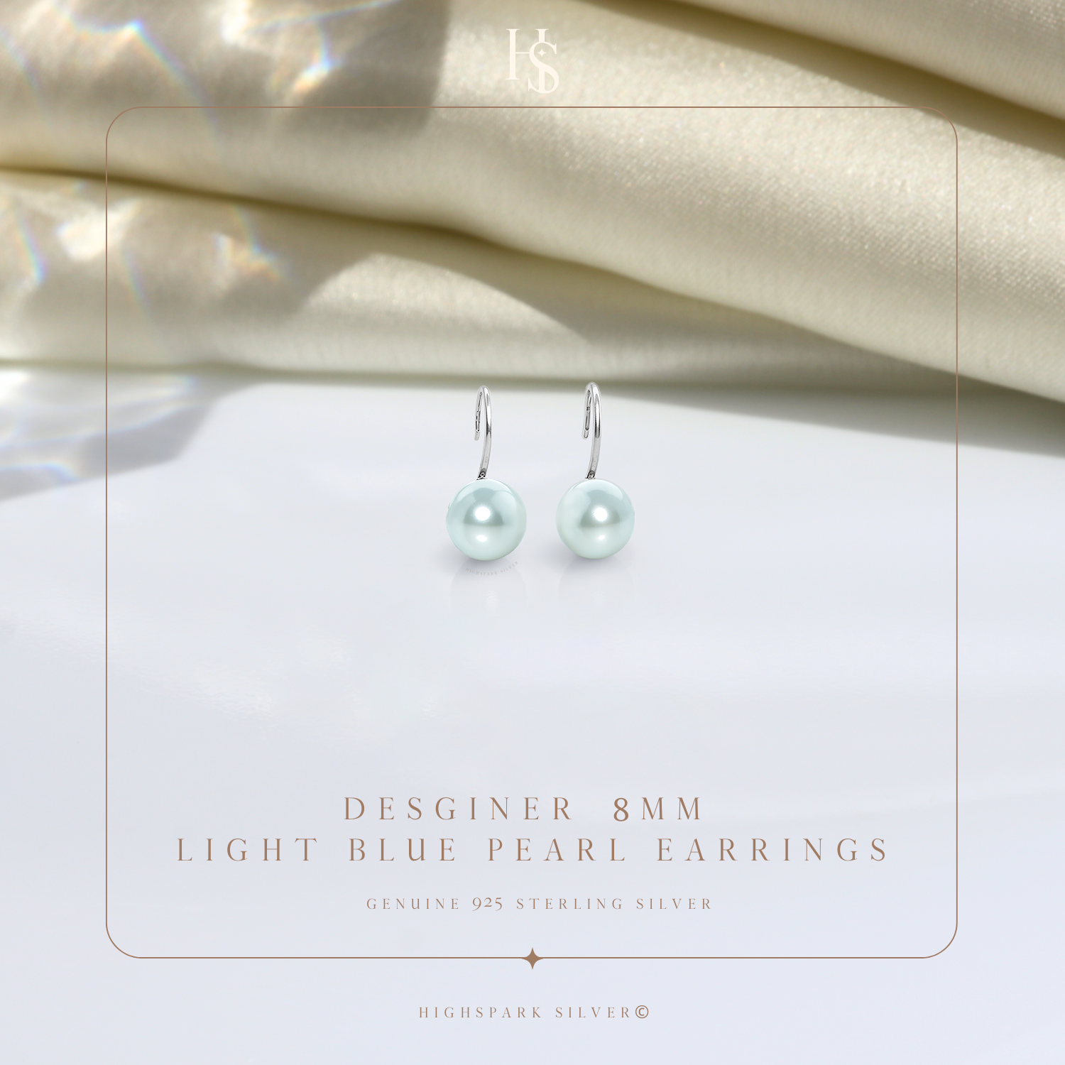 A pair of sterling silver hook earrings with round, light blue pearls, styled on a white surface with draped cream satin in the background.