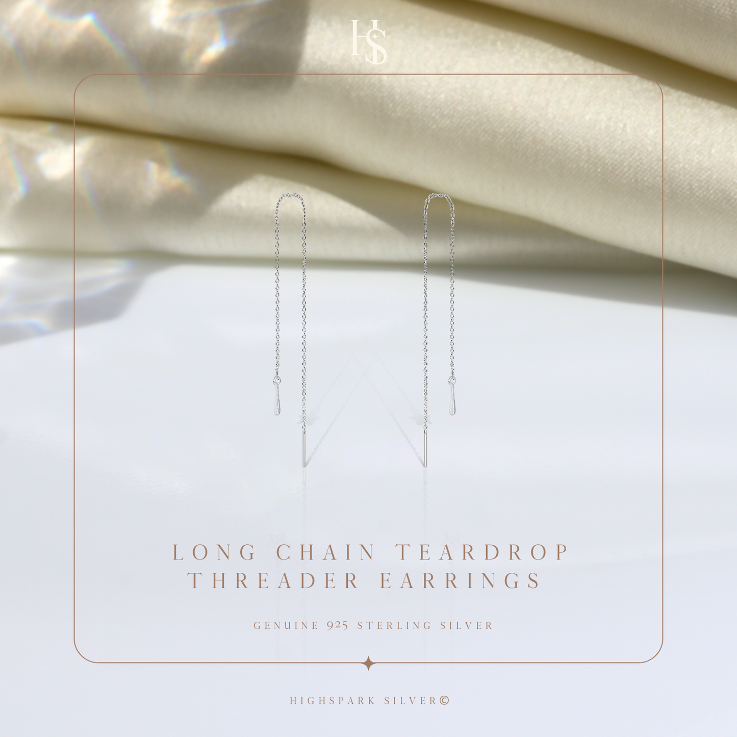 A pair of sterling silver threader earrings with a long chain and teardrop charm, styled on a white surface with draped cream fabric.