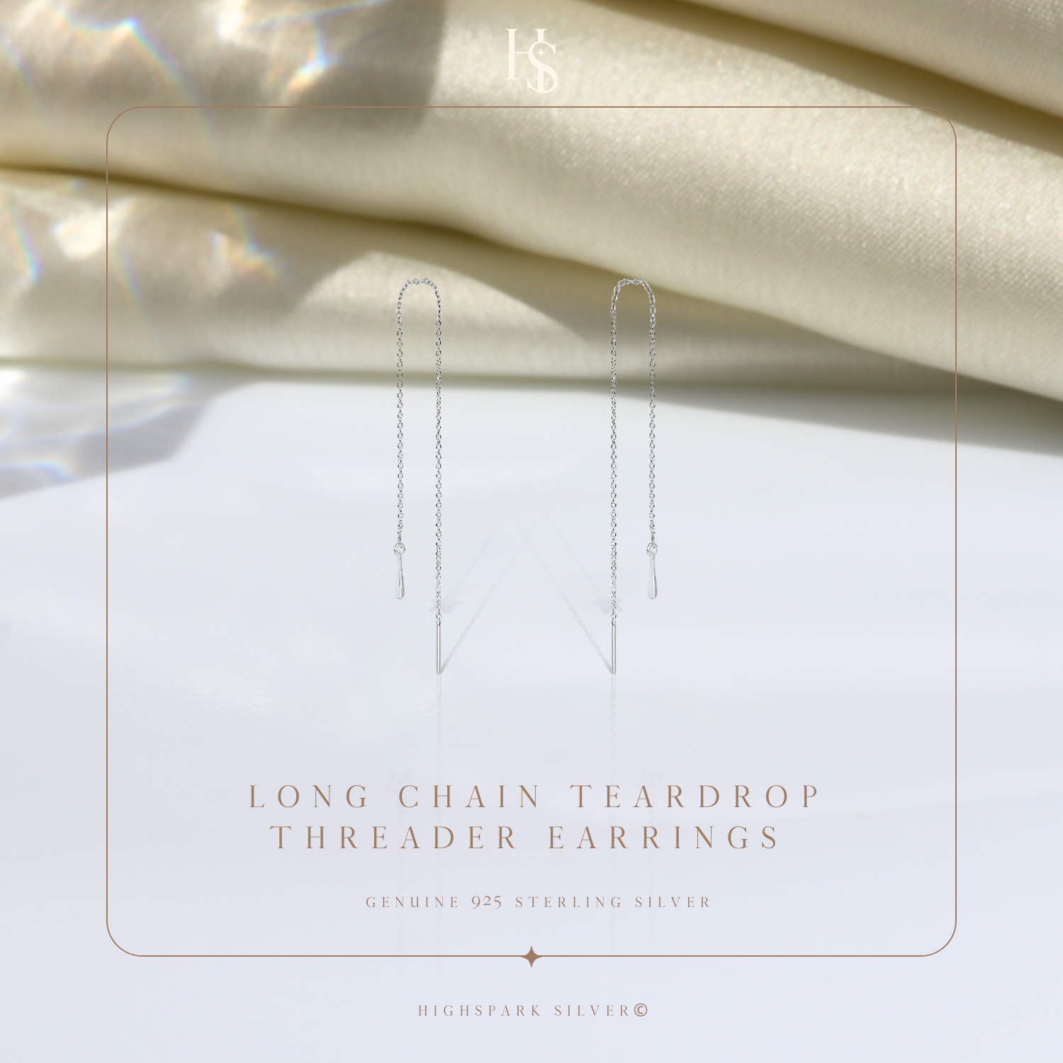 A pair of sterling silver threader earrings with a long chain and teardrop charm, styled on a white surface with draped cream fabric.