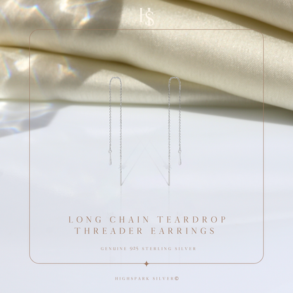 A pair of sterling silver threader earrings with a long chain and teardrop charm, styled on a white surface with draped cream fabric.