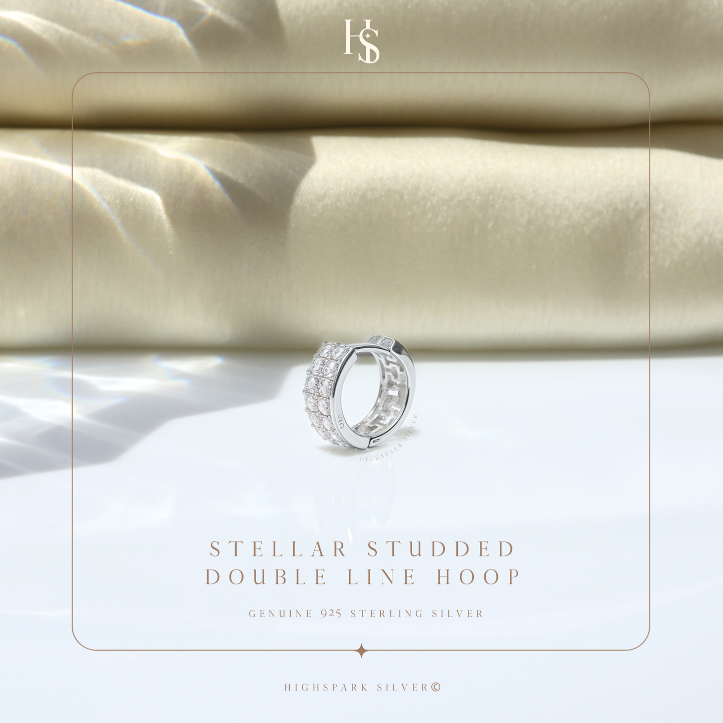 A sterling silver huggie hoop earring with two rows of pavé-set American diamonds, styled on a white surface with folded beige fabric.