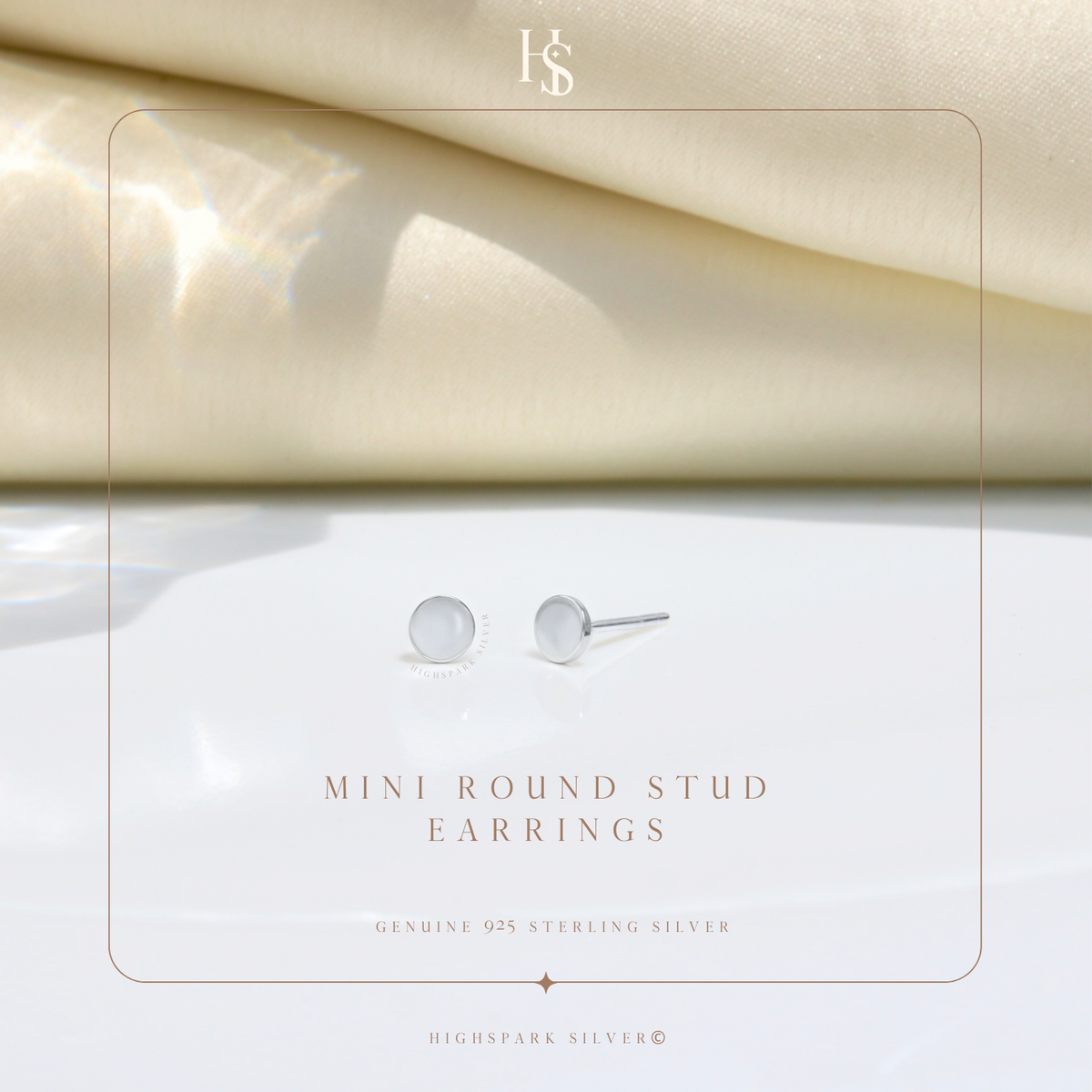 A pair of mini round stud earrings in sterling silver, styled on a white surface with draped cream fabric in the background.