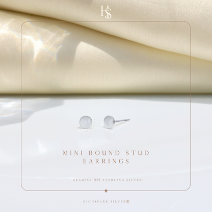 A pair of mini round stud earrings in sterling silver, styled on a white surface with draped cream fabric in the background.
