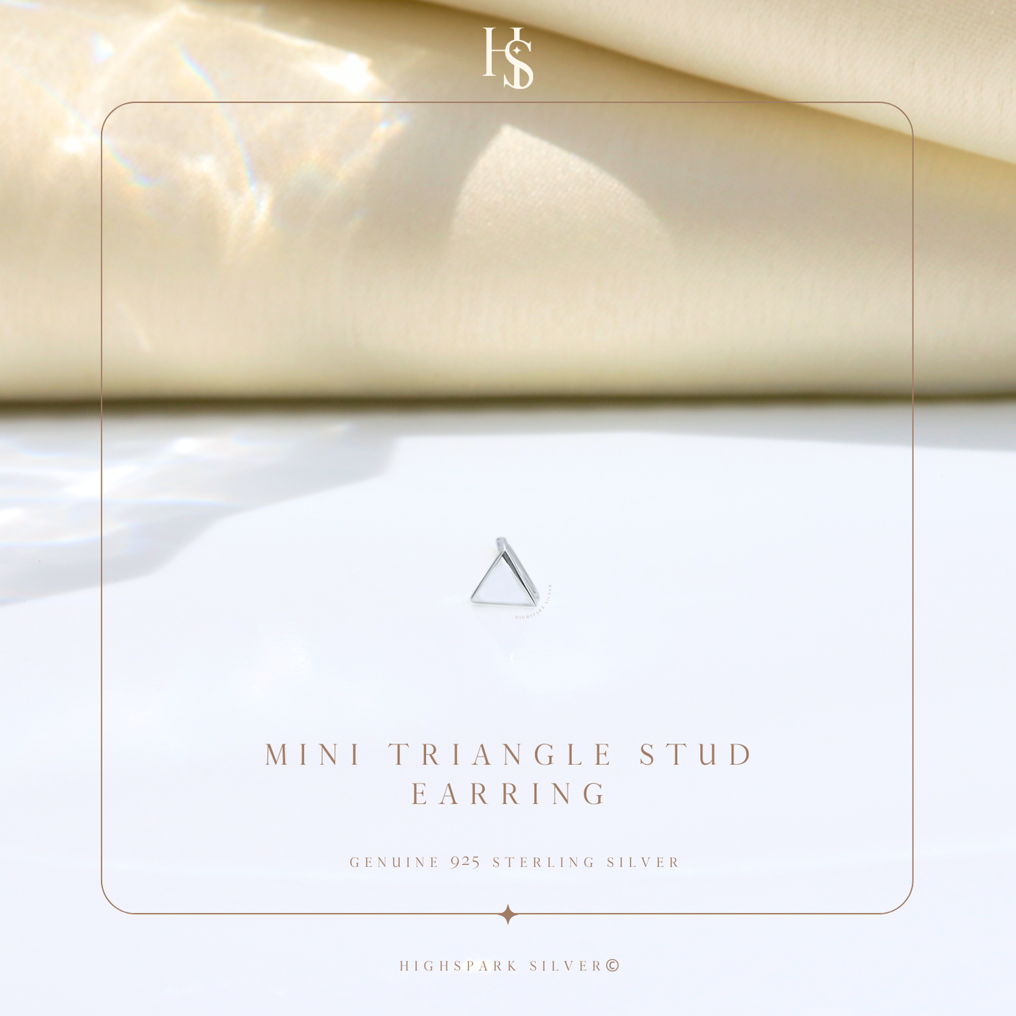 A single mini triangle stud earring in sterling silver, styled on a white surface with soft light and cream fabric in the background.