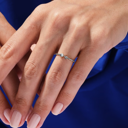 A minimalist sterling silver ring with a small round American diamond and a tiny bead cluster worn on the finger against a blue background.
