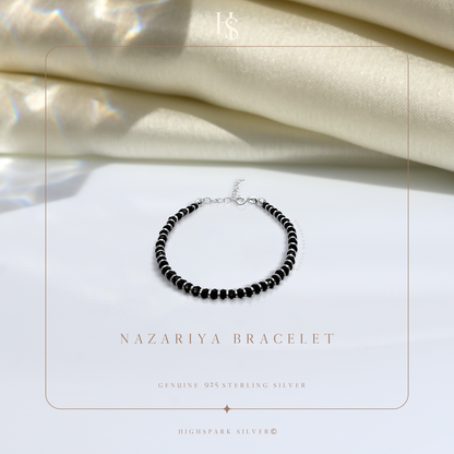 A nazariya bracelet with tiny black beads and sterling silver accents, styled on a white surface with draped ivory satin in the background.