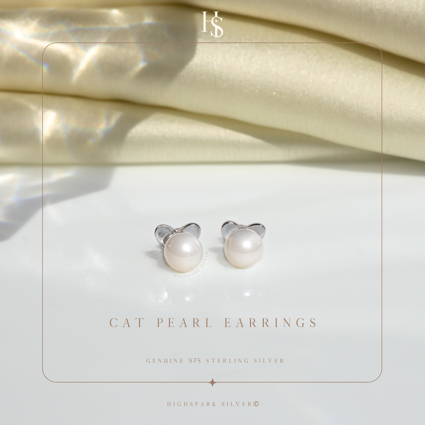 A pair of sterling silver cat stud earrings with freshwater pearls, styled on a white surface with folded cream satin in the background.