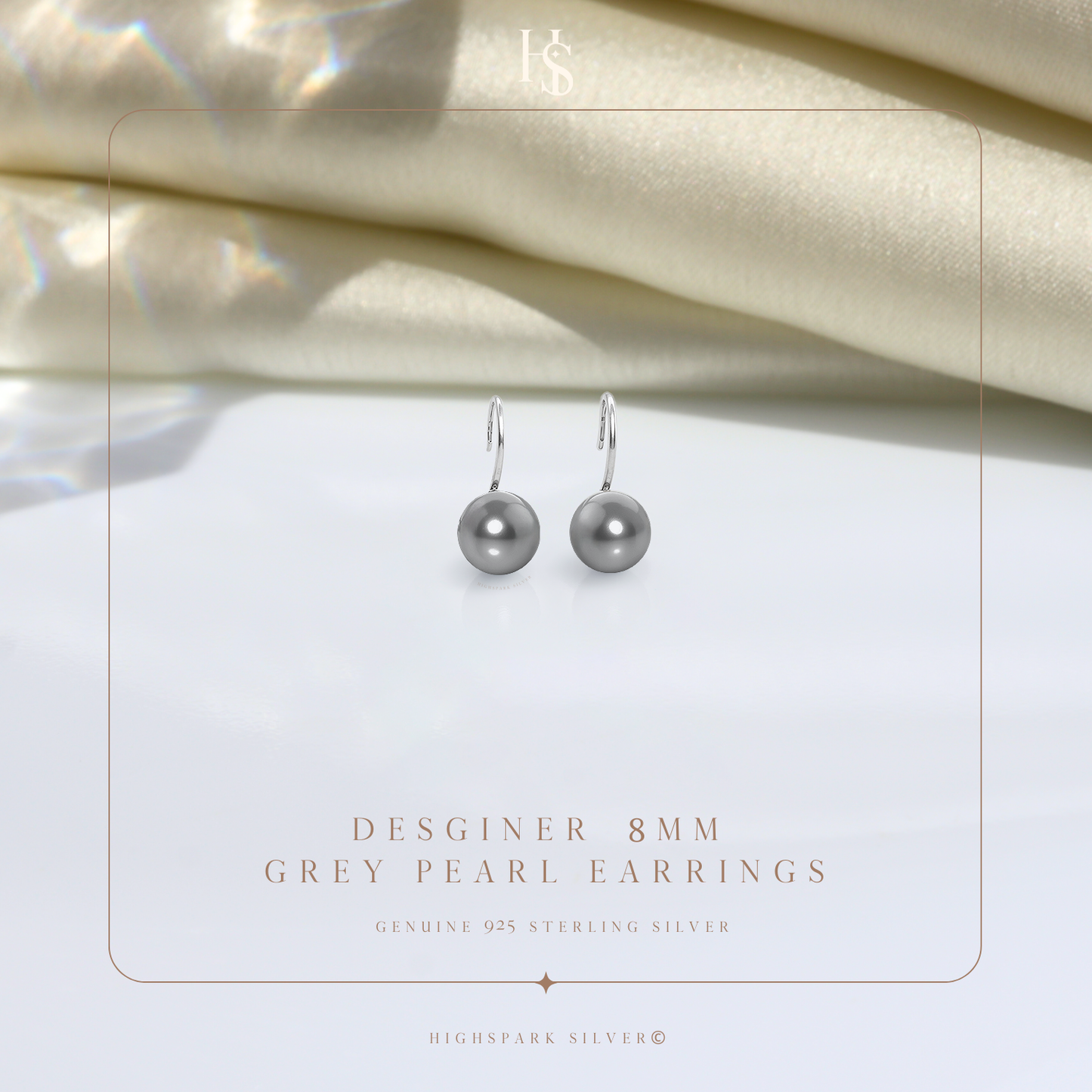 A pair of sterling silver hook earrings with round grey pearls, styled on a white surface with draped cream satin in the background.