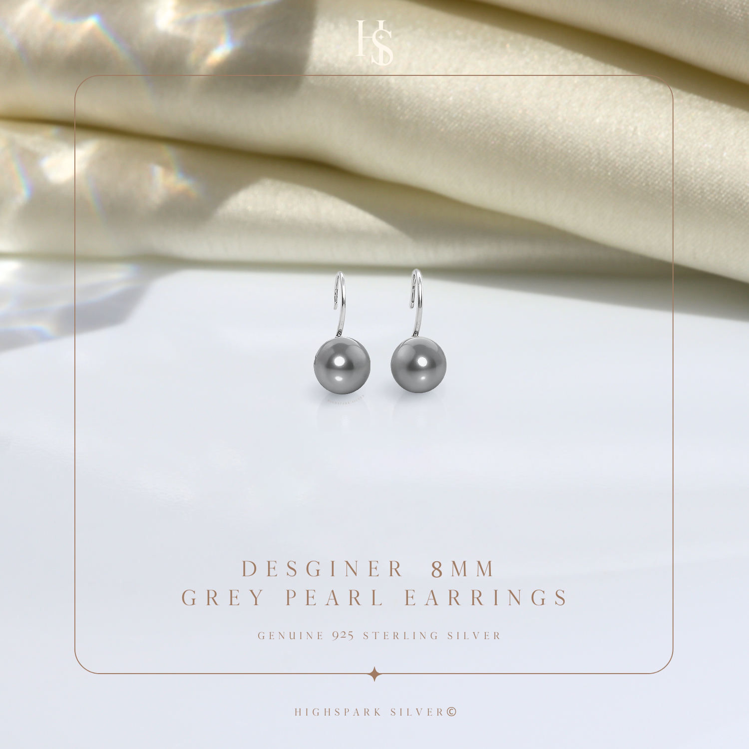 A pair of sterling silver hook earrings with round grey pearls, styled on a white surface with draped cream satin in the background.