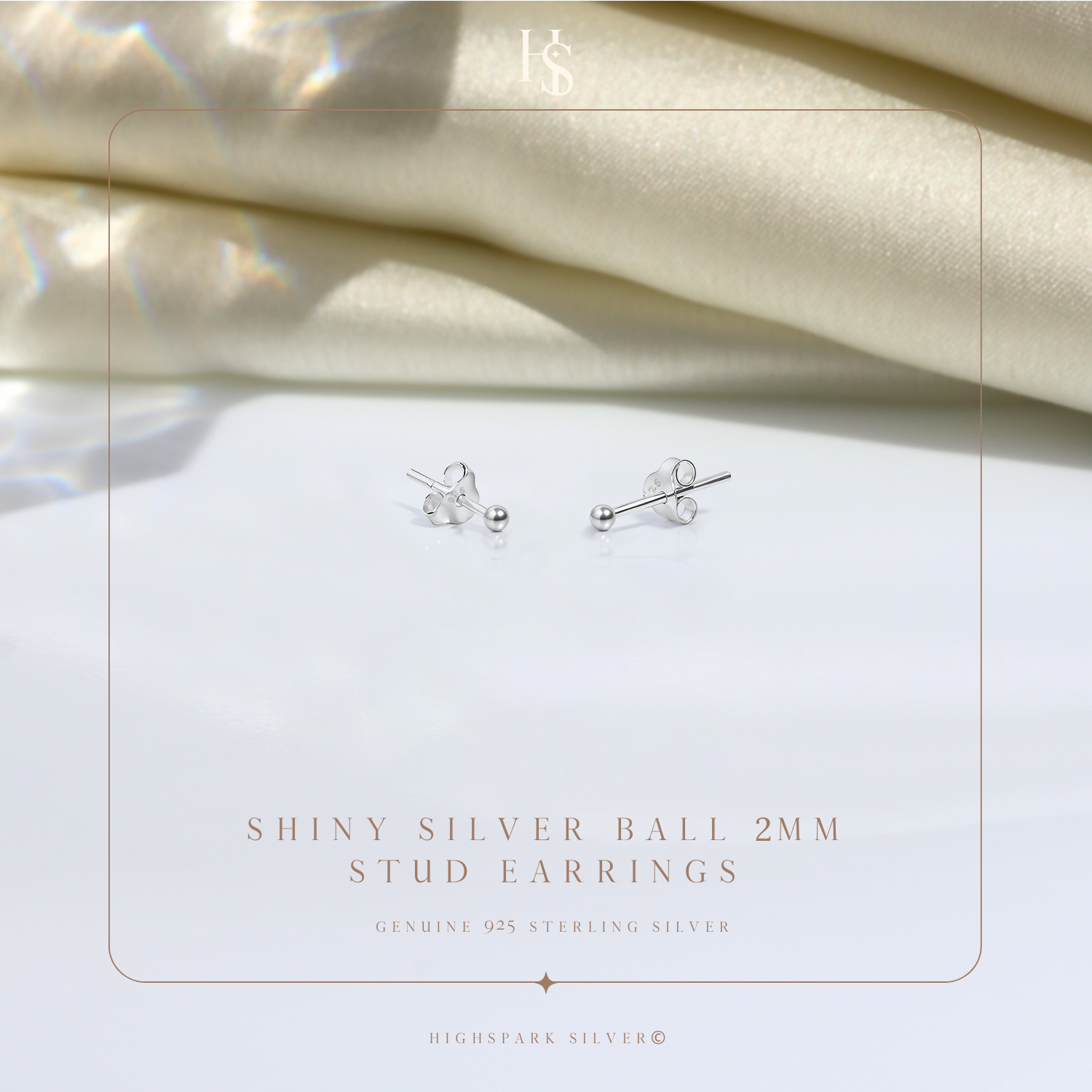 A pair of tiny ball stud earrings in sterling silver with push-back closures, styled on a white surface with draped cream satin fabric.