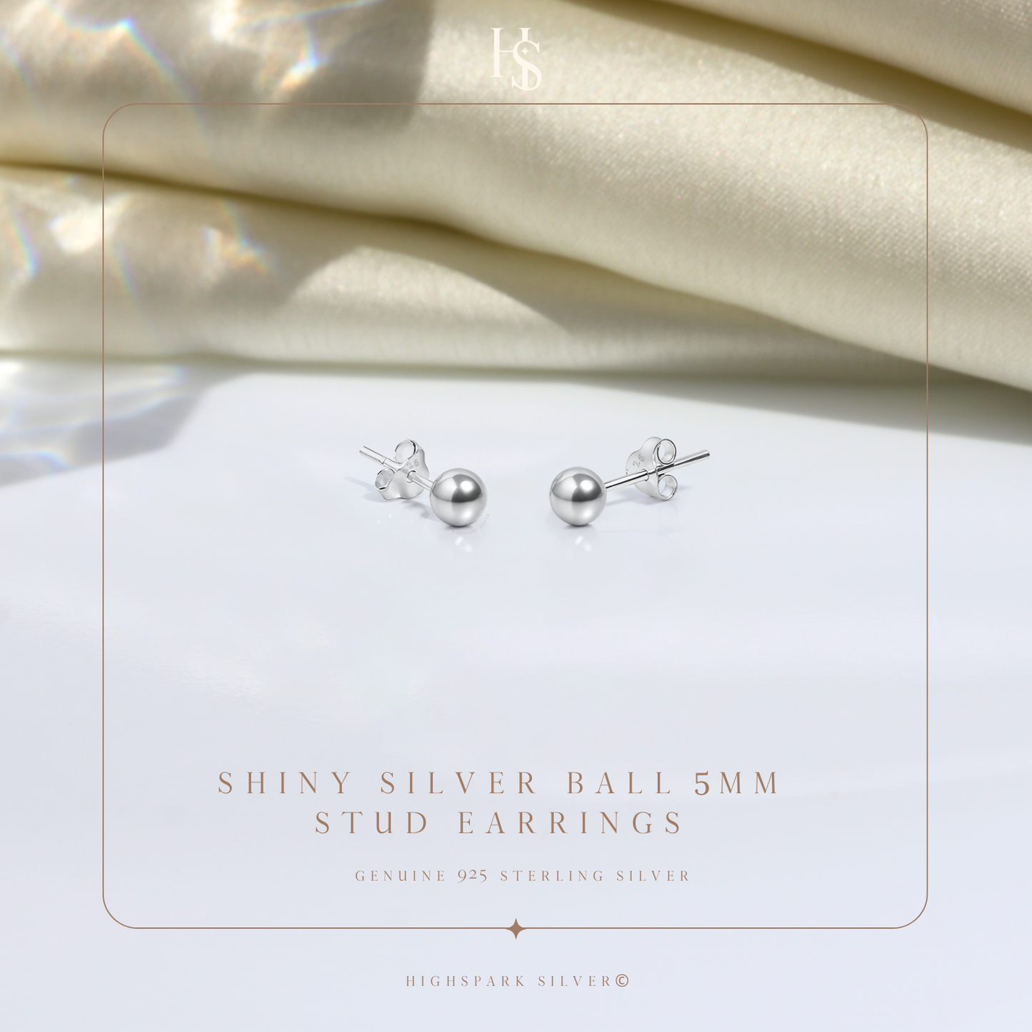 A pair of sterling silver ball stud earrings with butterfly backs, styled on a white surface with folded cream satin fabric behind.