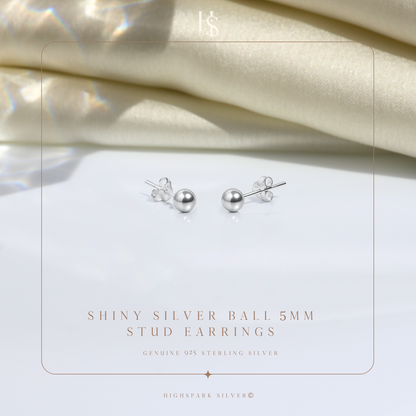 A pair of sterling silver ball stud earrings with butterfly backs, styled on a white surface with folded cream satin fabric behind.