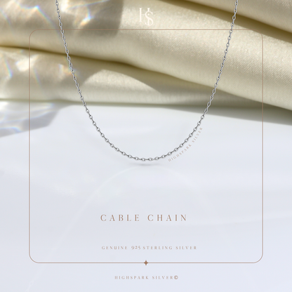 A simple sterling silver cable chain necklace styled on a white surface with draped cream satin fabric in the background.