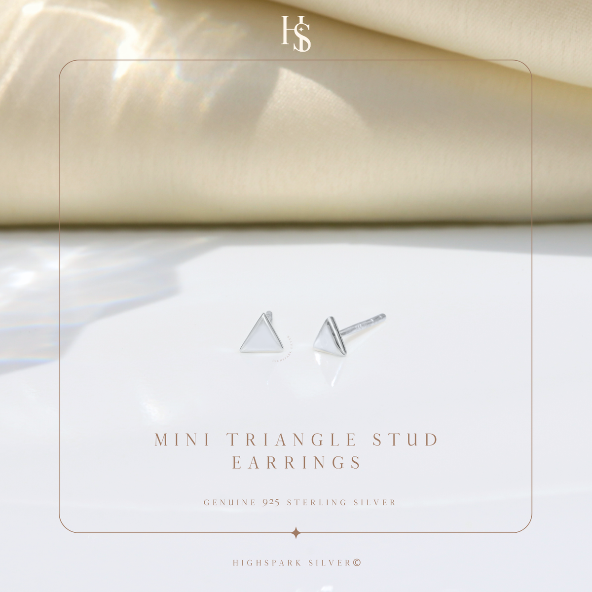 A pair of minimalist triangle stud earrings in sterling silver, styled on a white surface with a soft, cream-colored fabric background.