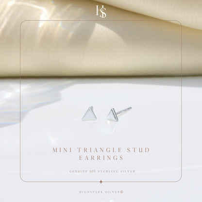 A pair of minimalist triangle stud earrings in sterling silver, styled on a white surface with a soft, cream-colored fabric background.
