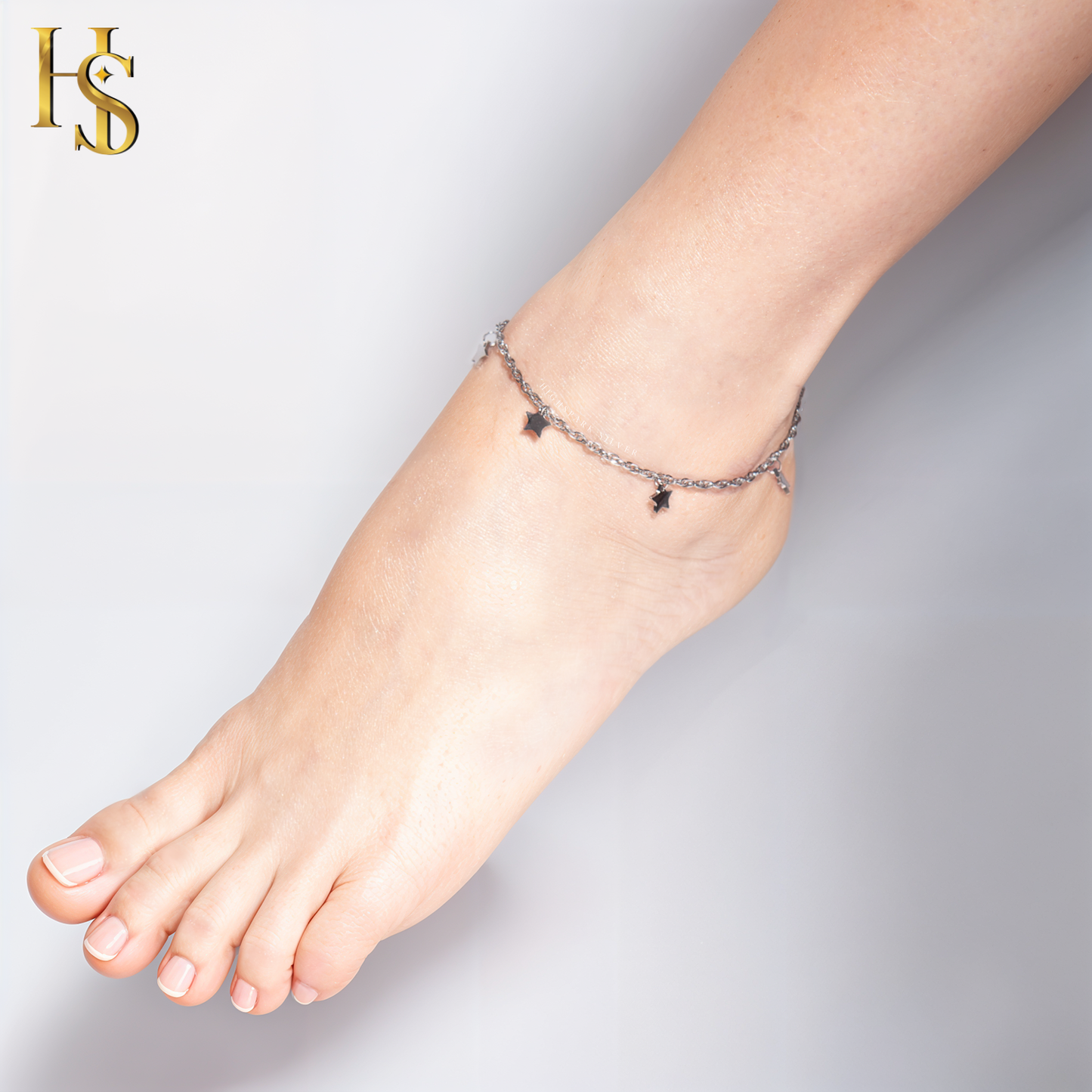 A sterling silver anklet with a delicate chain and small, dangling star charms is worn on the ankle against a plain background.