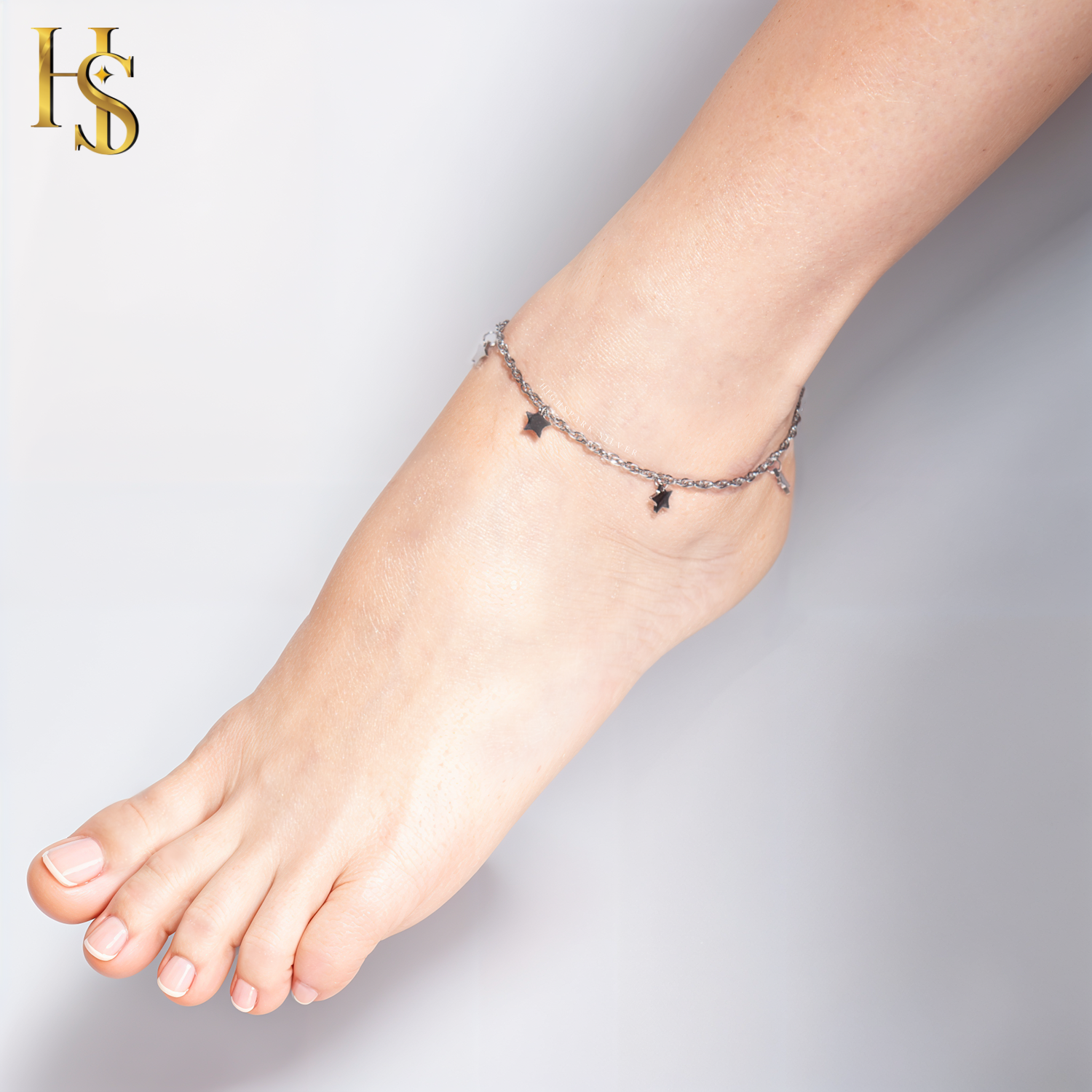 A sterling silver anklet with a delicate chain and small, dangling star charms is worn on the ankle against a plain background.