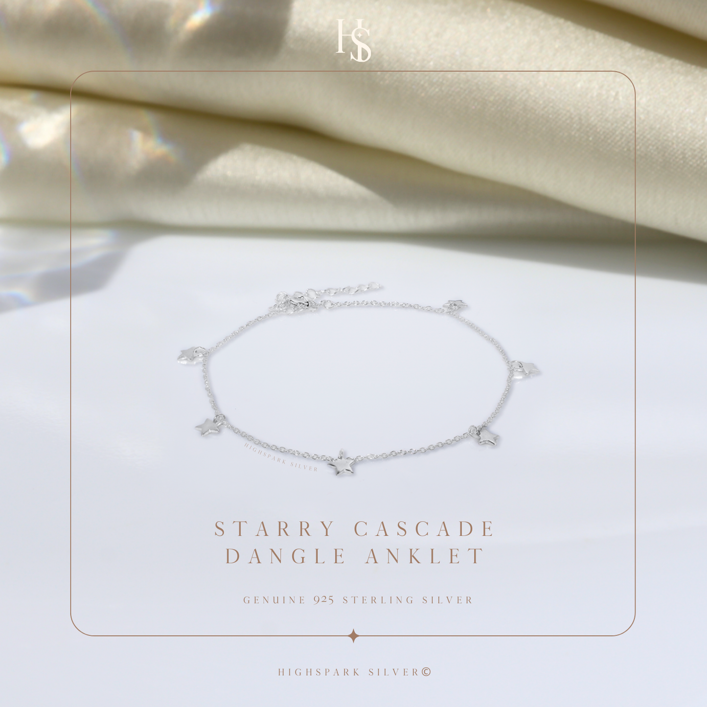 A sterling silver anklet with small dangling star charms, styled on a white surface with draped cream fabric in the background.