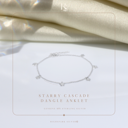 A sterling silver anklet with small dangling star charms, styled on a white surface with draped cream fabric in the background.