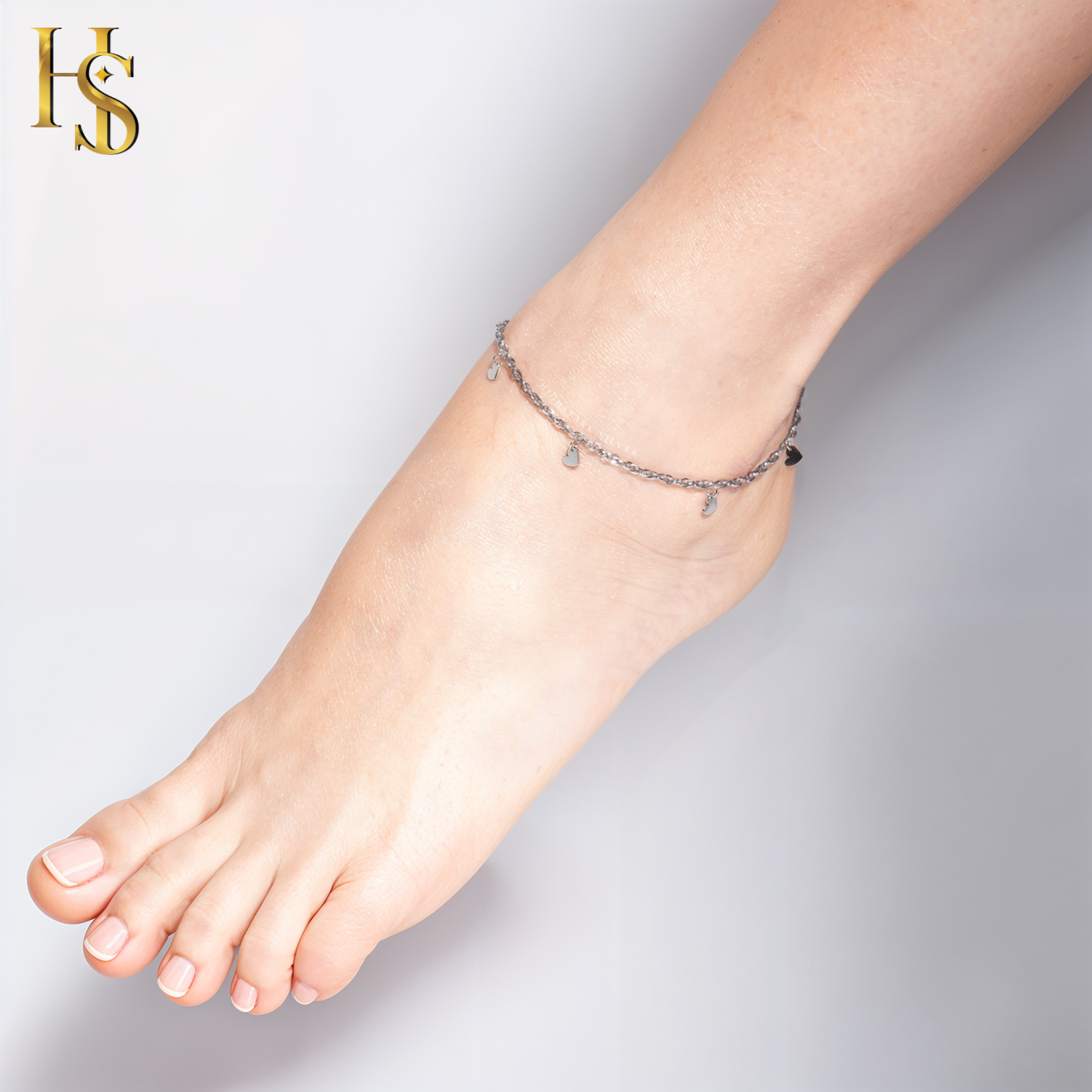 A sterling silver chain anklet with small, dangling heart charms is worn on a model's ankle against a plain background.