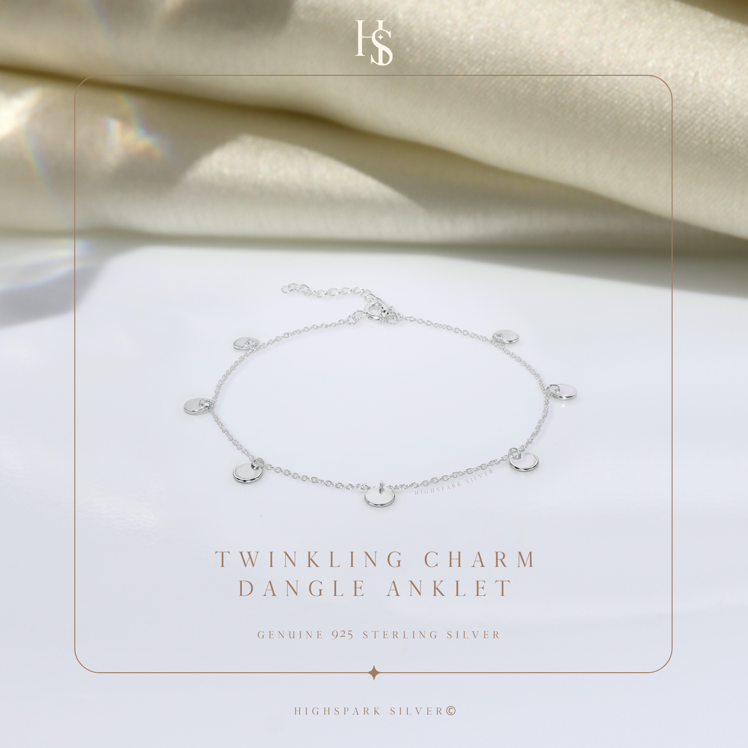 A sterling silver anklet with small, dangling disc charms, styled on a white surface with draped cream satin fabric in the background.