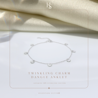 A sterling silver anklet with small, dangling disc charms, styled on a white surface with draped cream satin fabric in the background.