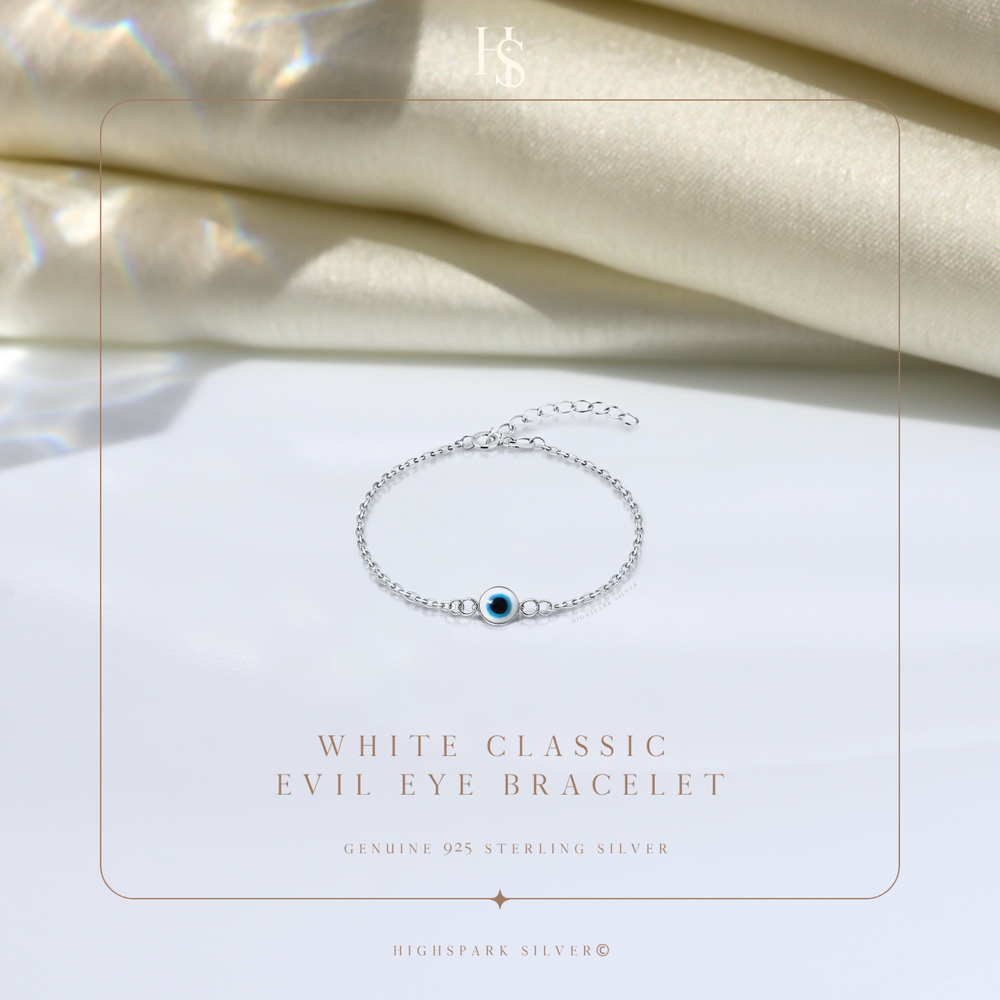 A sterling silver evil eye bracelet with a white and blue enamel charm, styled on a white surface with draped cream satin fabric.