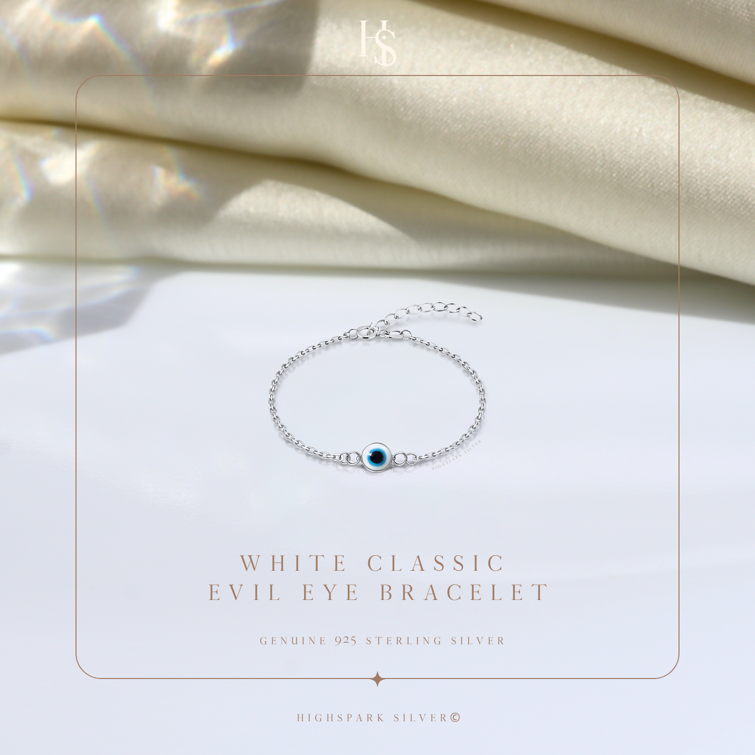 A sterling silver evil eye bracelet with a white and blue enamel charm, styled on a white surface with draped cream satin fabric.
